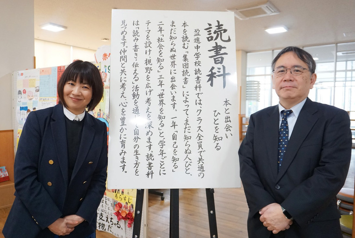 図書室に掲げられている読書科の理念と一緒に写る上山朋子教諭と中馬淳常務理事